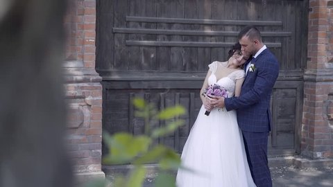 pretty bride in long white dress with purple bouquet leans on handsome husband against wooden fence slow motion
