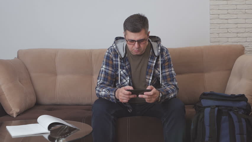 A middle-aged man sitting on a sofa in a hotel room or apartment uses the Internet in a smartphone, next to him is a large tourist backpack.