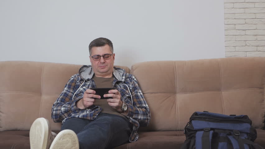 A middle-aged man sitting on a sofa in a hotel room or apartment uses the Internet in a smartphone, next to him is a large tourist backpack.
