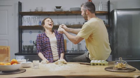 Caucasian boyfriend and girlfriend having fun in kitchen, throwing flour and dough in each other and laughing.