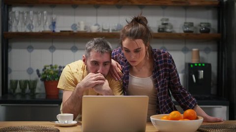 Couple in kitchen tensely watching sports match on computer, then suddenly reacting excitedly and feeling extremely happy
