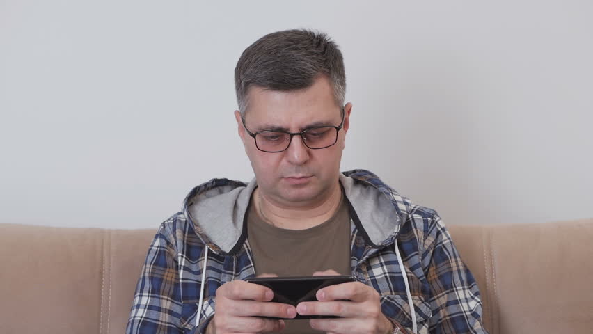 A middle aged man in glasses is using mobile internet in a smartphone while sitting on a sofa in a room.
