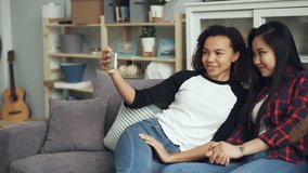 Smiling young women Asian and African American making online video call looking at smartphone screen talking and laughing sitting on couch in nice apartment.