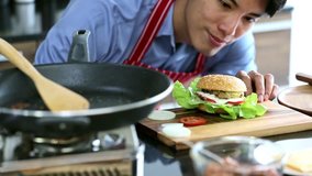 Slow motion scene video of Asian man in red apron sitting, he is smiling and feeling proud of his hamburger.
