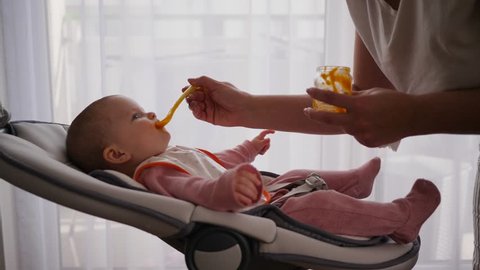 Mother leans to the child that lie in baby swing and feed with first food