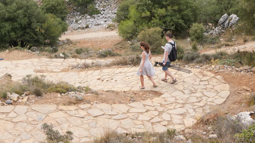 Young woman and man walk down by winding stone paved trail, view from above. Tourist couple come back from Psychro Cave