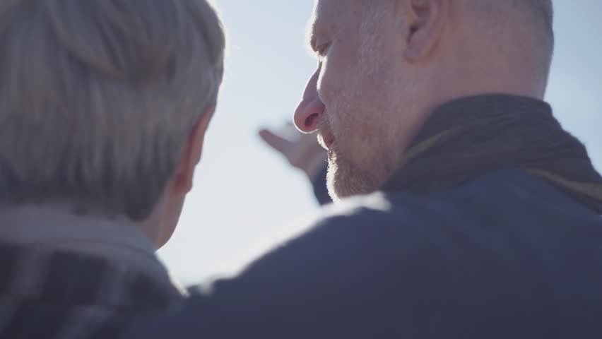 Back of woman with grey short hair and back of bald man standing outdoors. Mature married couple communicating in the sun outdoors. Family relationships.