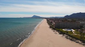 Flying over the coastline in Denia, Spain with some touristic apartments. The beach is desert, small waves on the sea, Montgo and Segaria mountains in the background