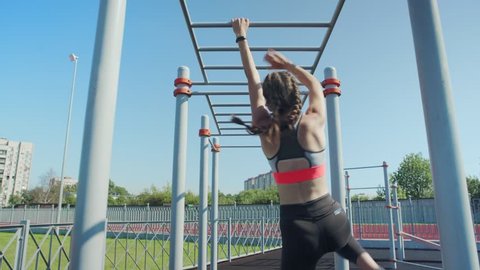 Back view of athletic teenage girl doing monkey bars climbing exercise and recovering after it at street workout spot, fitness tracker on her wrist