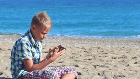 Cute white kid using smartphone while sitting on sand of summer sunny beach. Real time full hd video footage.