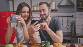 Happy lovely couple having breakfast and using smartphones while sitting together on kitchen