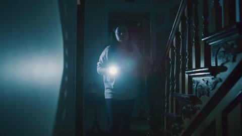 A woman with a flashlight walks downstairs in a dark house at nighttime. 
