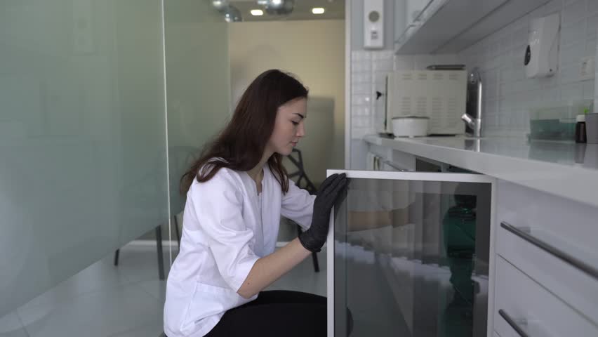 Close-up scene of a young pretty female dentist who is getting a dental instruments in a metal boxes out of a closet. Woman is absolutely confident and calm about her work.