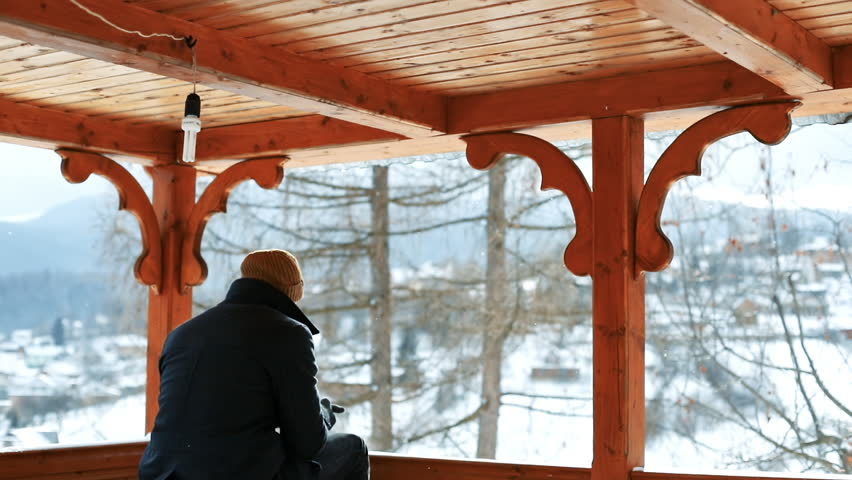 Back view of inspired guy delighting amazing nature on winter day. Man sitting inside a wooden arbor against beautiful countryside between snowy mountains.