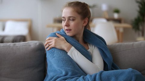 Sick  young woman feeling cold fever freezing no central heating problem at home covering with warm plaid blanket shivering sitting on couch got flu influenza virus grippe symptoms concept