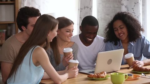 Happy young diverse friends having fun watching comedy movie, online tv show or funny social media video on laptop, multiracial millennial people students group laughing use computer together in cafe
