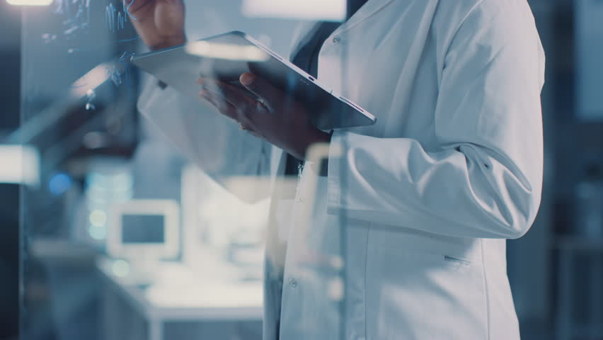 In the Research Laboratory Smart and Beautiful African American Female Scientist Wearing White Coat and Protective Glasses Writes Formula on Glass Whiteboard, References Her Tablet Computer Royalty-Free Stock Footage #1025738195