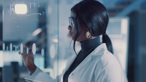 In the Research Laboratory Smart and Beautiful African American Female Scientist Wearing White Coat and Protective Glasses Writes Formula on Glass Whiteboard, References Her Tablet Computer