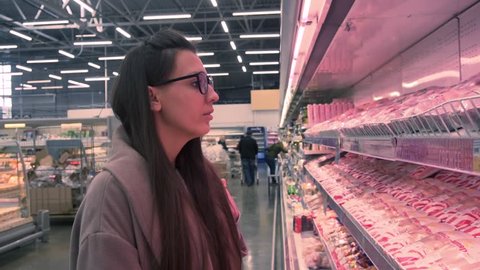 Young woman with glasses and a coat in a supermarket chooses packages of chicken meat. Overall plan.