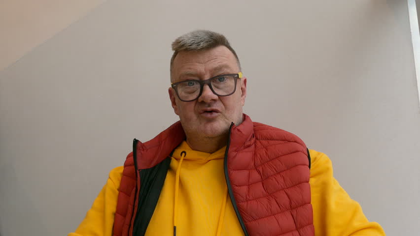 Elderly man of caucasian nationality, in bright clothes, drinking coffee from white cup, waiting for ordered food, sitting in restaurant or cafe.