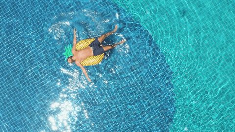 Drone aerial view of Young man floating on inflatable pineapple over blue swimming pool enjoying sunbathing and vacations in tropical destination. People travel tourism holidays concept 
