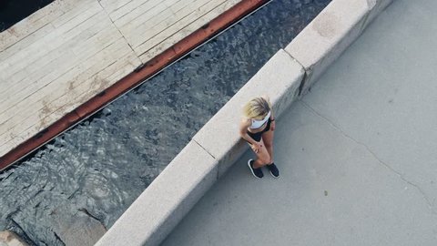 Directly above view of blonde athletic young woman leaning on embankment parapet near river while resting after jogging and checking results on fitness tracker on her wrist