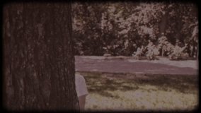 Family video archive. Retro camera 8 mm. Old film. A blonde boy peeks out from behind a tree, playing hide-and-seek on a sunny summer day