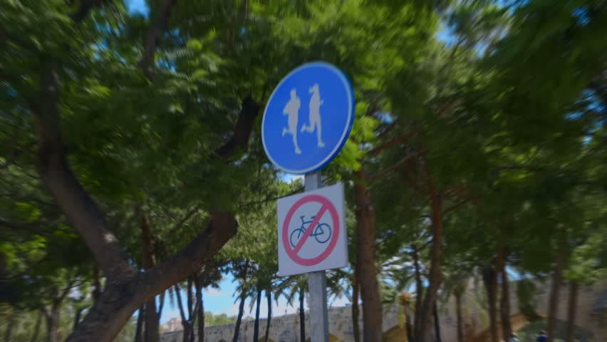 In the park, a blue road sign allowing runners to run and prohibiting cycling in red against the background of trees and sun flares into the camera lens. Shot in motion