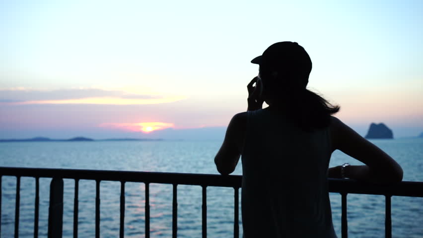 Silhouette rear view of asian woman wear cap and talking with someone by mobile phone during on the ferry boat with seascape background while sunset time.