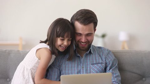 Happy family dad and cute child daughter watching cartoons on laptop at home on sofa, father with kid girl having fun looking at computer screen enjoying funny video or playing online game together
