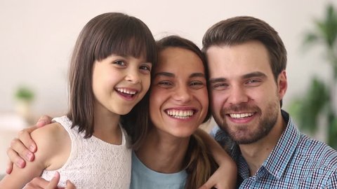 Happy family of three mom dad and cute child little daughter embracing laughing looking at camera, smiling young parents with kid girl having fun hug enjoy sincere communication warm relationships