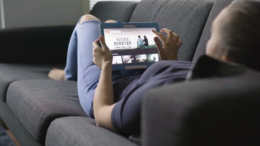 Woman using movie stream service with tablet. Choosing series or film and lying on couch in home living room. Watching an episode of a tv show with mobile device. Streaming on demand video. Royalty-Free Stock Footage #1025769953