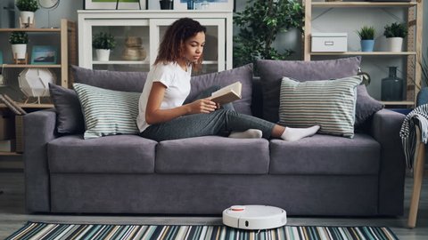 Good-looking African American student is turning on robotic hoover and reading book enjoying hobby and relaxing on couch while gadget is cleaning floor.
