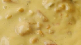 Very close video of stirring hot chicken corn chowder with a wood spoon while heating illuminated with natural lighting.