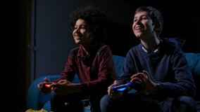 Diverse teen friends holding joysticks, playing video game console, sitting on couch late evening. Two smiling boys in casual clothing communicating while playing home console, using game controllers