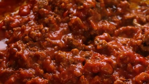Close slow motion video of marinara sauce being stirred into browned beef and cheddar cheese hamburger meat in a pan while cooking with a wood spoon illuminated with natural lighting.