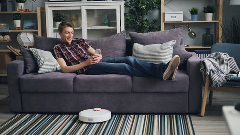 Happy student is using smartphone looking at screen and smiling relaxing on comfortable couch while robotic vacuum cleaner is vacuuming carpet and floor.