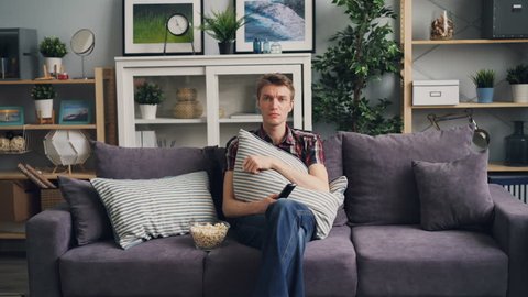 Zoom-in of sensitive guy watching TV and crying over tragic film sitting on couch and holding remote control and pillow during leisure time at home. People and emotions concept.