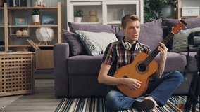 Young man successful vlogger is recording video playing the guitar and singing sitting on floor at home and using camera. Blogging and apartment concept.