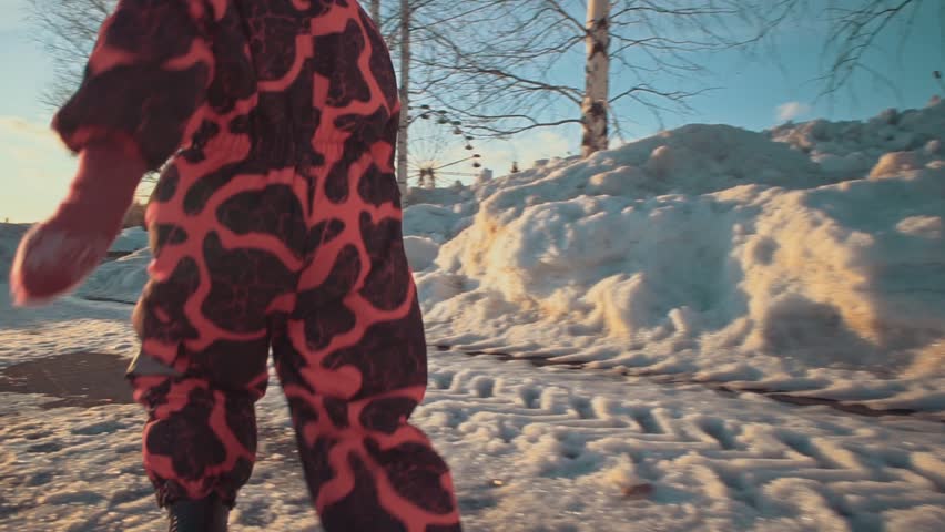 Happy child flees on the paths in the park. A small girl in a pink jumpsuit runs through the snow on a clear sunny evening at sunset, slow motion.