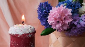 Looped video: close-up of burning candle, made in the shape of Easter cake, stands next to a wicker basket with beautiful multi-colored hyacinths.
