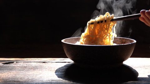 Slow motion Chopsticks to tasty noodles with steam and smoke in bowl on wooden background, selective focus., Asian meal on a table, junk food concept