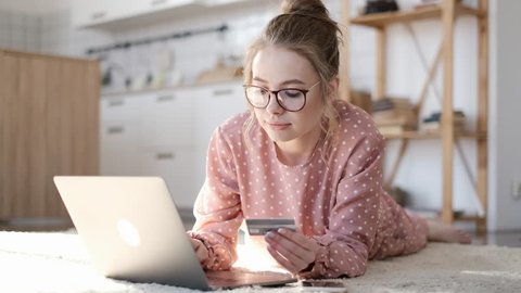 Smiling woman with laptop and credit card at home. Girl with laptop and bank card indoors. Beautiful woman lying in the living room shopping online with credit card. Easy pay using digital gadget.