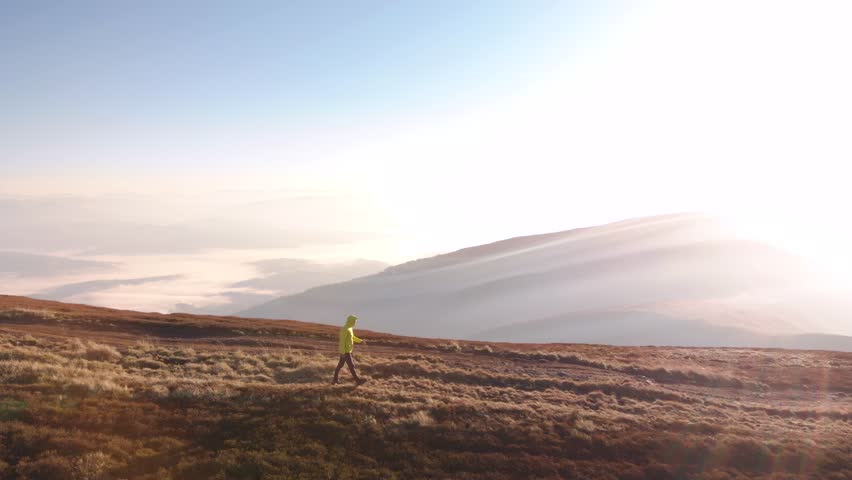 Aerial drone flying slider like shot against sun. Man hiker walking on mountain road with distant valley on horizon at sunrise