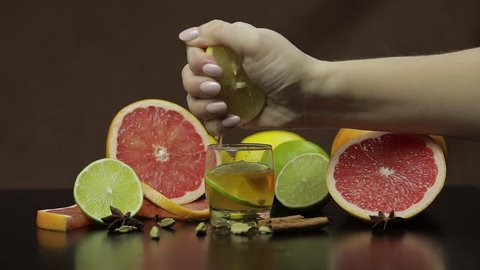 Woman squeezes juice from half of lemon fruit into a glass cup with alcohol cocktail. Fresh fruits in the background. Lime Grapefruit. Prepare a cocktail of whiskey, cognac or liqueur. Slow motion
