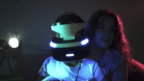 Mother hugging son wearing VR headset, boy holding joystick and push the buttons with young woman while playing video game , family spending time together in dark room illuminated neon lights