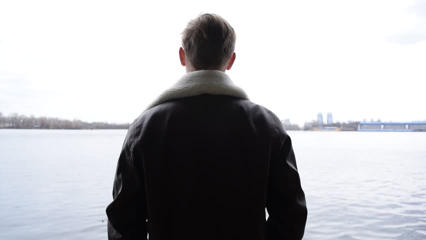 A young guy in a leather jacket shoots with a gun near the river in the city
