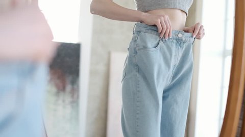 Happy female in old jeans after successful diet. Young woman dancing in front of a mirror rejoicing weight loss before the summer season. Healthy food and sports