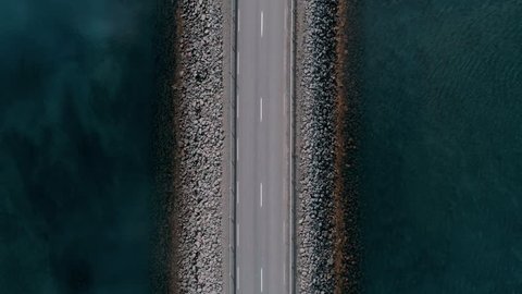 Straight down drone footage of bridge or road in middle of canal or lake separating sea from sweet water. Commuter or tourist traffic vehicles and cars drive by. Icelandic landscape
