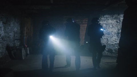 Special ops military SWAT team members walking in dark musty underground tunnel with flashlights approaching captured police officer. Wide to medium close shot on a RED camera.